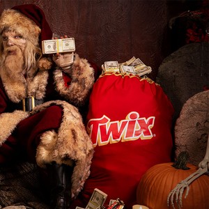 Zombie Claus holding cash beside a giant TWIX sack and pumpkins